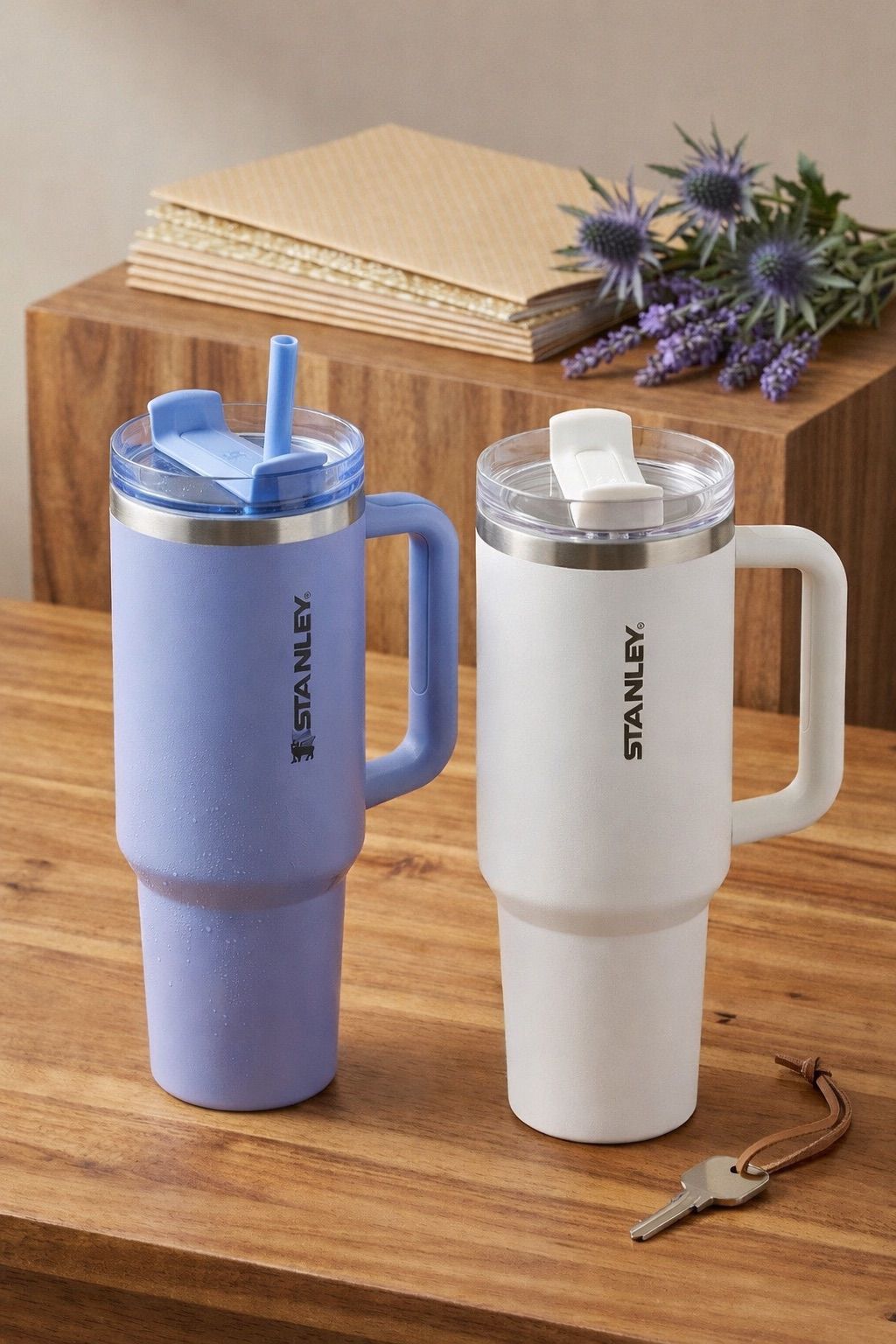 Two Stanley travel mugs, one purple and one white, on a wooden surface with a book and flowers in the background.